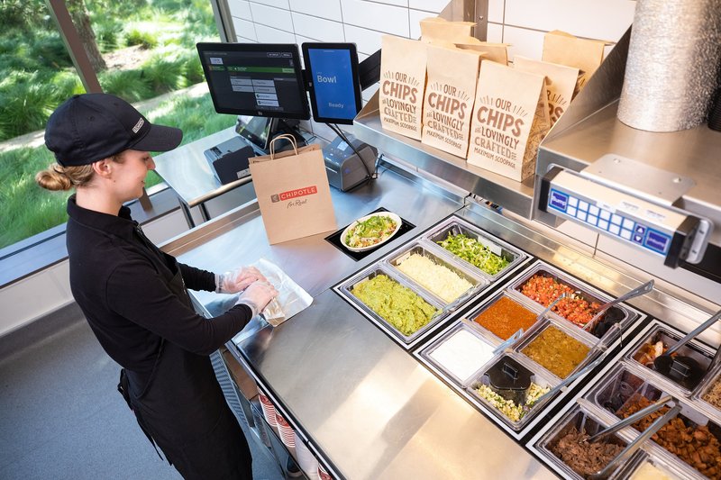 Chipotle Augmented Makeline by Hyphen: a worker at a counter with ingredient pans while bowls are assembled automatically below